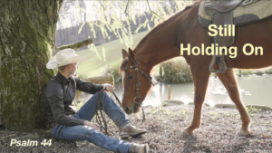 Cowboy leaning against a tree holding the reins of a horse with the text "Still Holding On - Psalm 44"